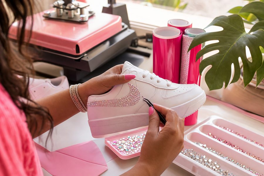 Rhinestones being applied by hand to a white sneaker beside rolls of pink and turquoise transfer vinyl